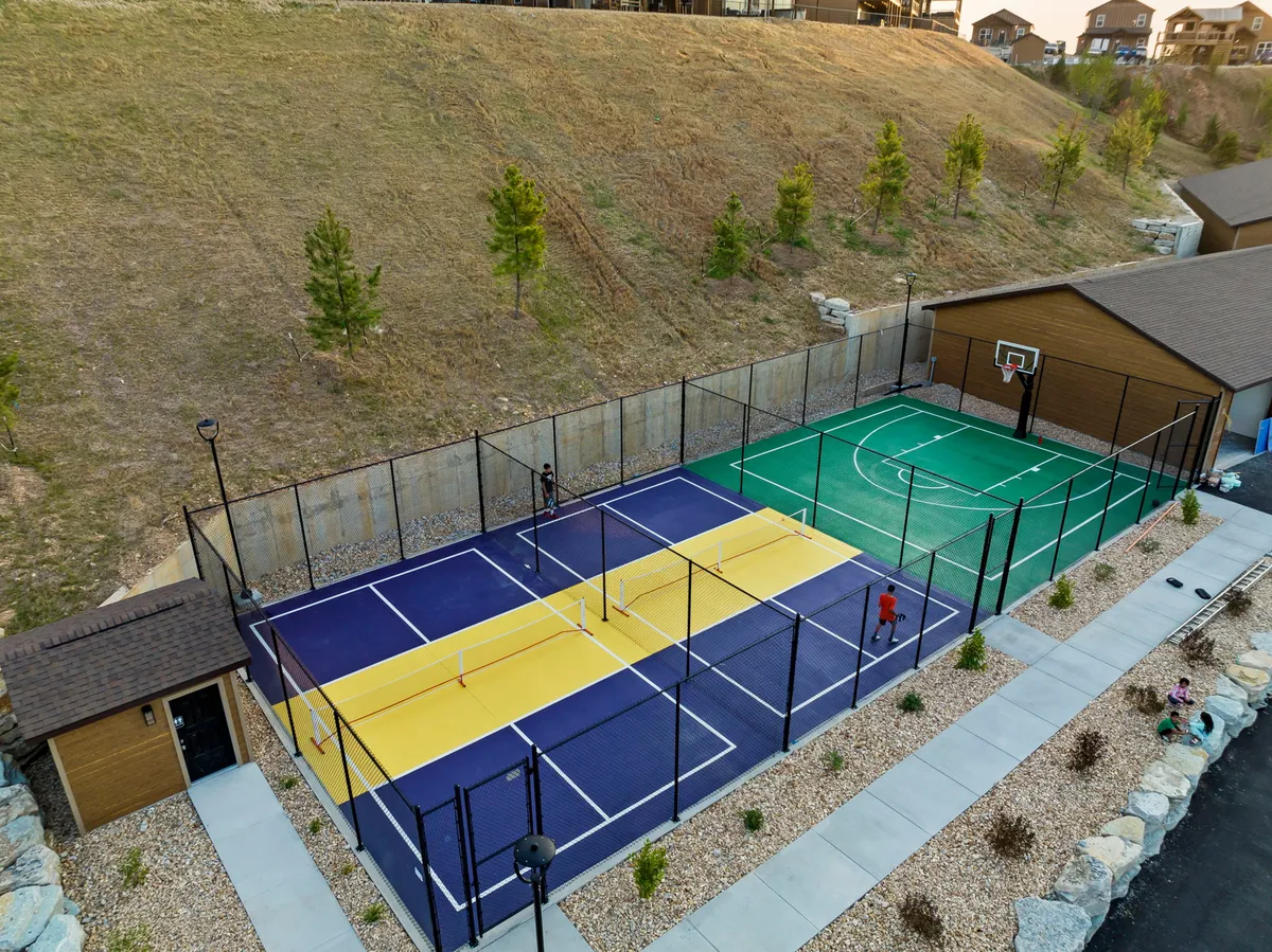 An exterior multi-sport court boasts a basketball hoop, lined for pickleball, surrounded by lush greenery and accessible pathways. The area invites friendly competitions and active play among friends and families.