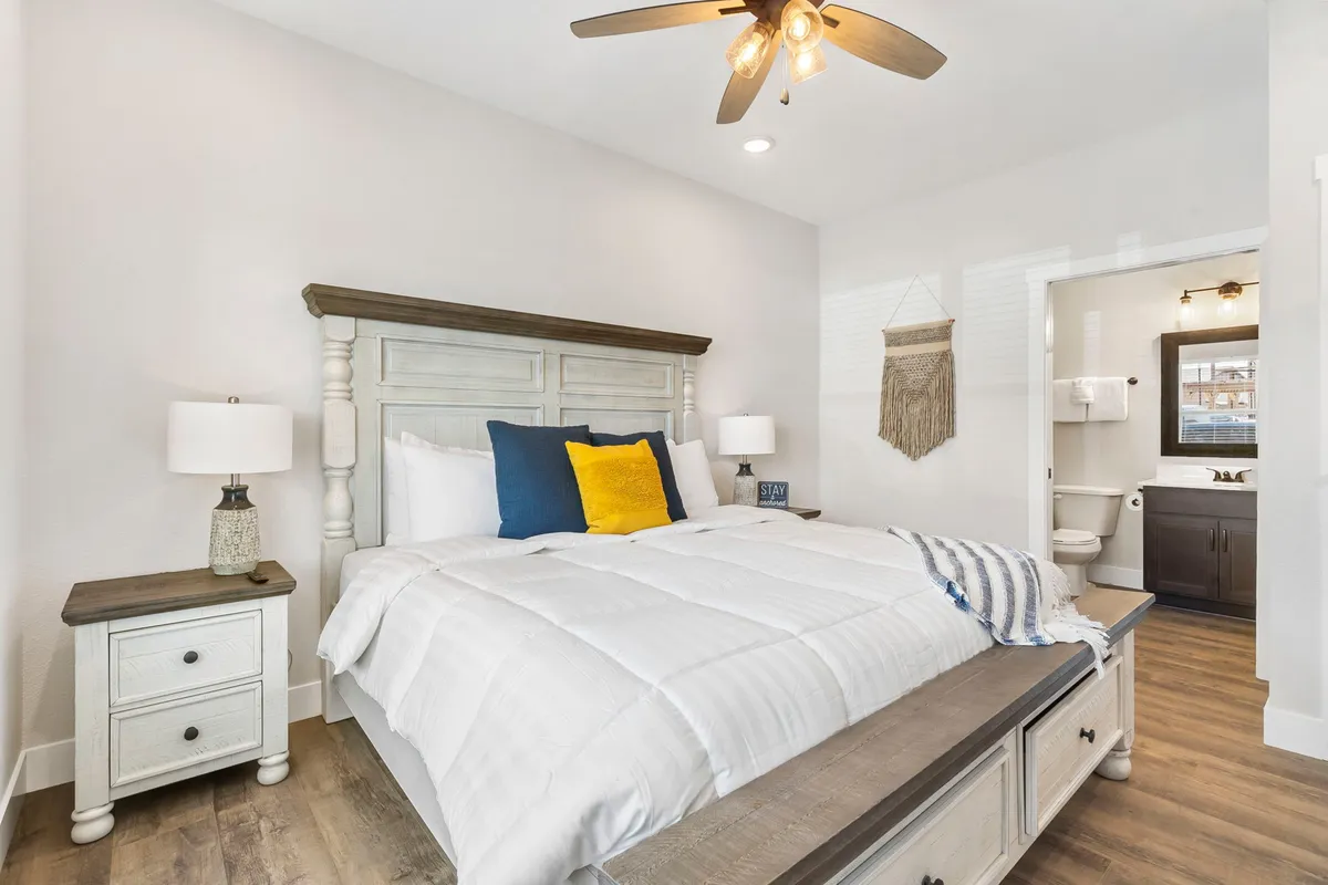 A tastefully decorated king bedroom featuring a plush bed with decorative pillows, nightstands with matching lamps on either side, a wooden ceiling fan overhead, and an open door revealing a private ensuite bathroom. The room exudes warmth and comfort, inviting guests to relax and unwind.