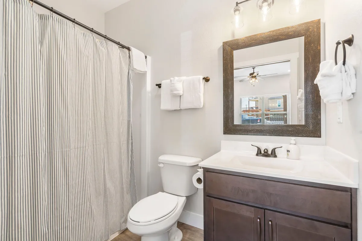 A bright, modern ensuite bathroom featuring a vanity with a sink, a large framed mirror, a striped shower curtain, and a toilet. The clean design and natural light suggest a refreshing and private space for self-care.