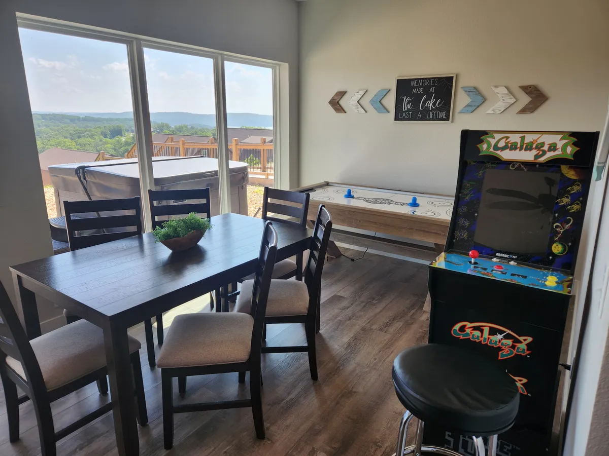 A cozy indoor space with a dining table adjacent to an arcade game, large sliding windows, and a decorative wall sign, bathed in natural light. The space is ideal for both social gatherings and casual games.