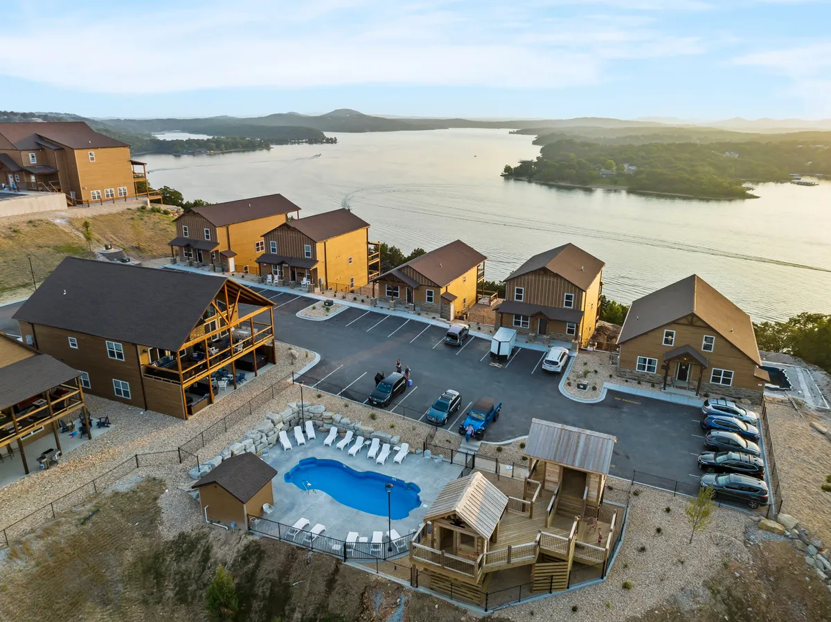 A vibrant resort scene with a swimming pool, playground structure, a row of cabins, and a sweeping lake view. Families can be seen swimming, sunbathing, and gathering, with the resort amenities and communal atmosphere taking center stage.