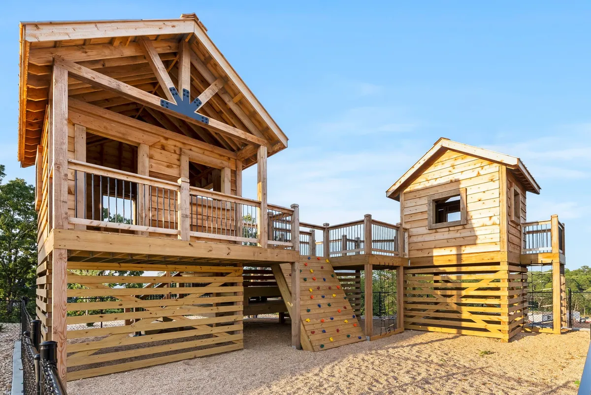 This joyful exterior image features a playhouse structure nestled among trees, complete with climbing stairs and a slide. Children can be seen actively playing, climbing, and socializing in the natural setting, embodying imaginative play and physical activity in a family-friendly environment.