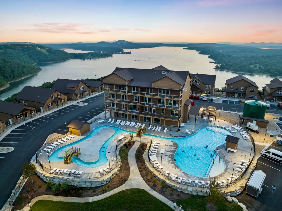 An inviting exterior shot captures the resort's swimming pools with the hotel building and scenic lake in the background. Guests are seen swimming, sunbathing, and relaxing by the pool while taking in the tranquil landscape, including the sparkling water and surrounding hills.